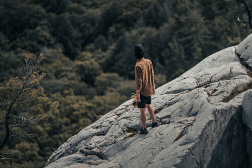 man-in-hat-stands-on-the-edge-of-a-rock-formation-overlooking-forest