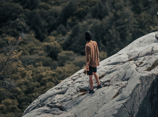 man-in-hat-stands-on-the-edge-of-a-rock-formation-overlooking-forest
