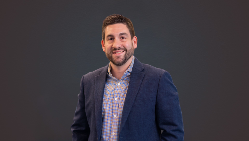 Smiling man in a navy blazer and checkered shirt posing against a dark wall.
